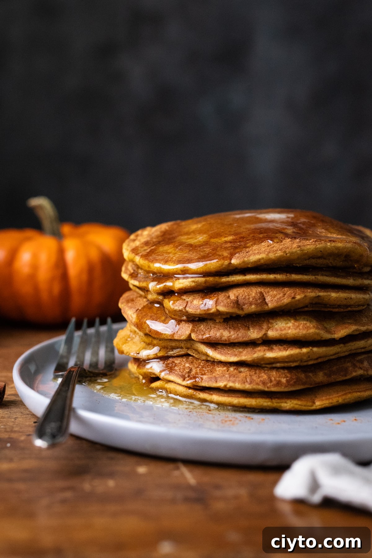 A stack of pumpkin spice pancakes on a gray plate with a fork laying next to the pancakes and an orange pumpkin in the background.