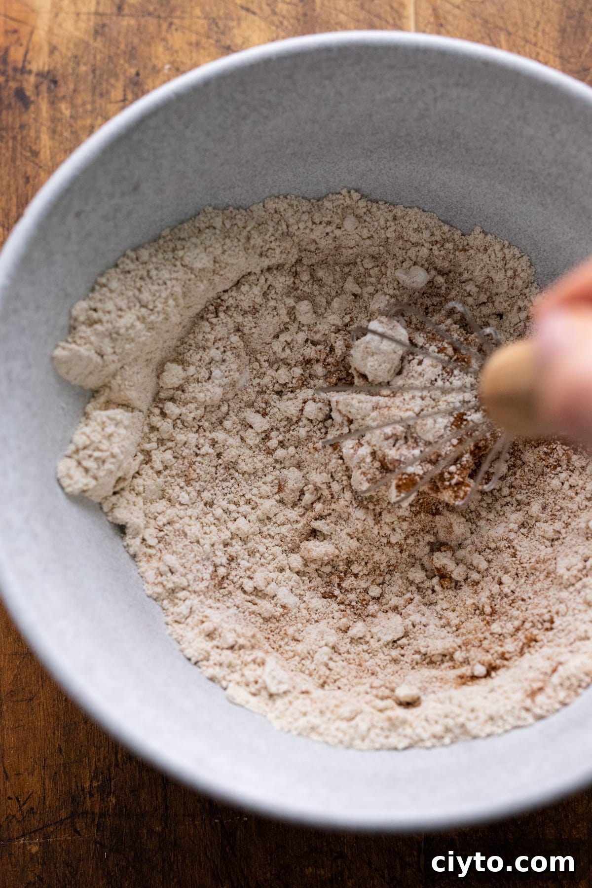 A hand using a whisk to stir dry ingredients for pancakes in a white bowl.