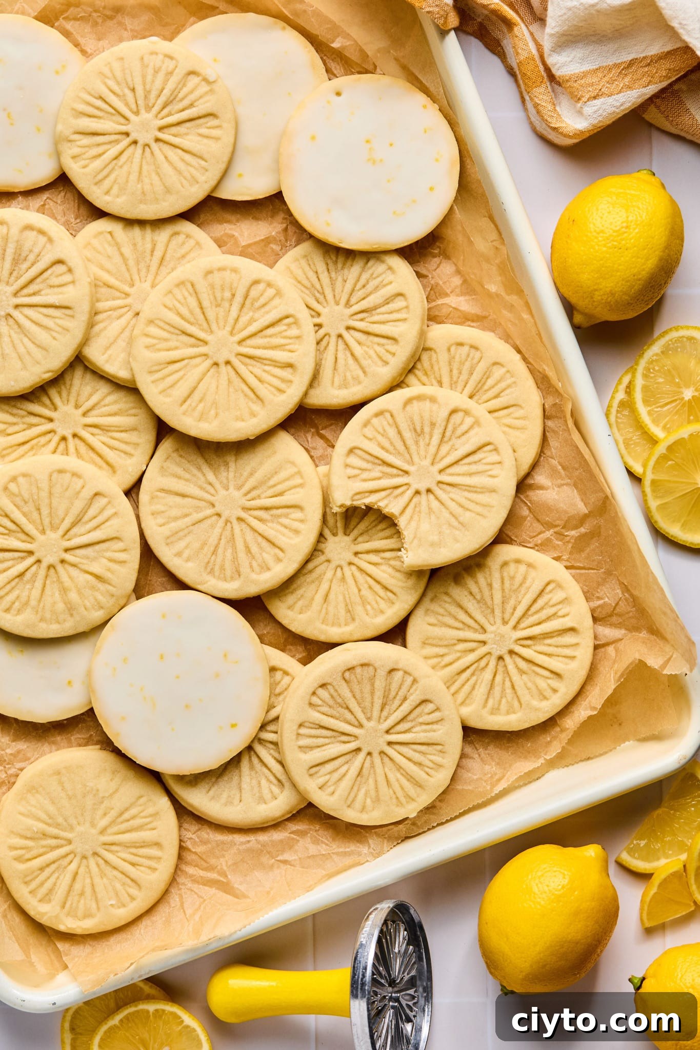 Picture of lemonade cookies on parchment-lined baking sheet. One cookie has a bite taken out of it. There are lemons and the metal lemon stamp surrounding the baking sheet.