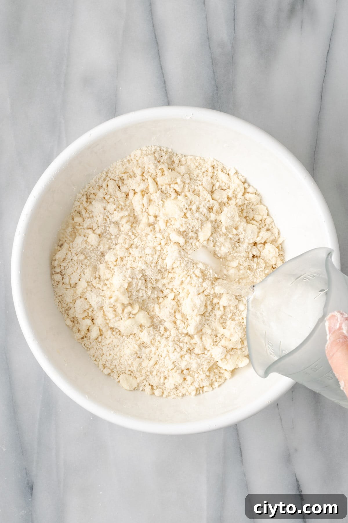 How to Make a Flaky Gluten-Free Pie Crust 11 A measuring cup of water being poured over the partially mixed pie dough.