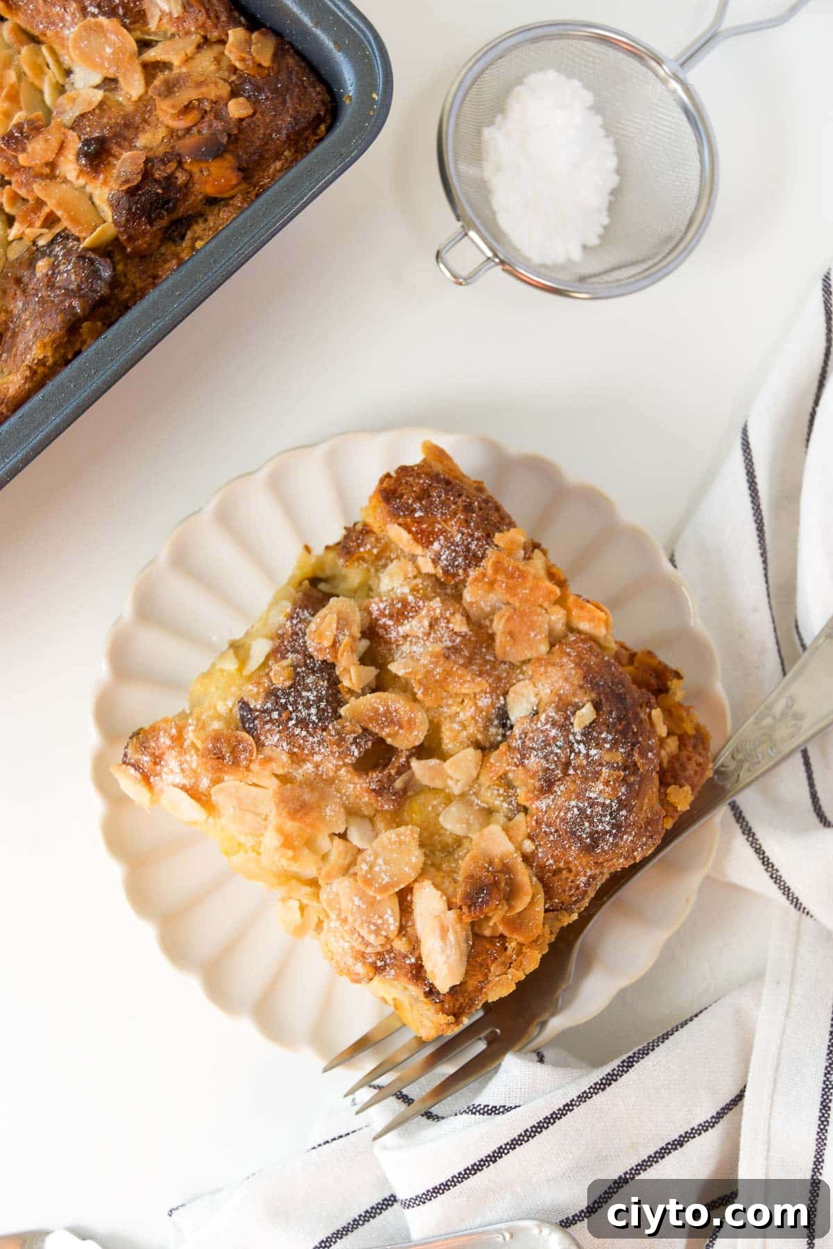 a slice of panettone bread pudding on a white scalloped plate with a fork next to a square pan of more bread pudding