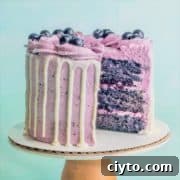 A blueberry cake on a pedestal with a slice taken out showing layers of moist blueberry cake, blueberry buttercream, and blueberry compote filling.