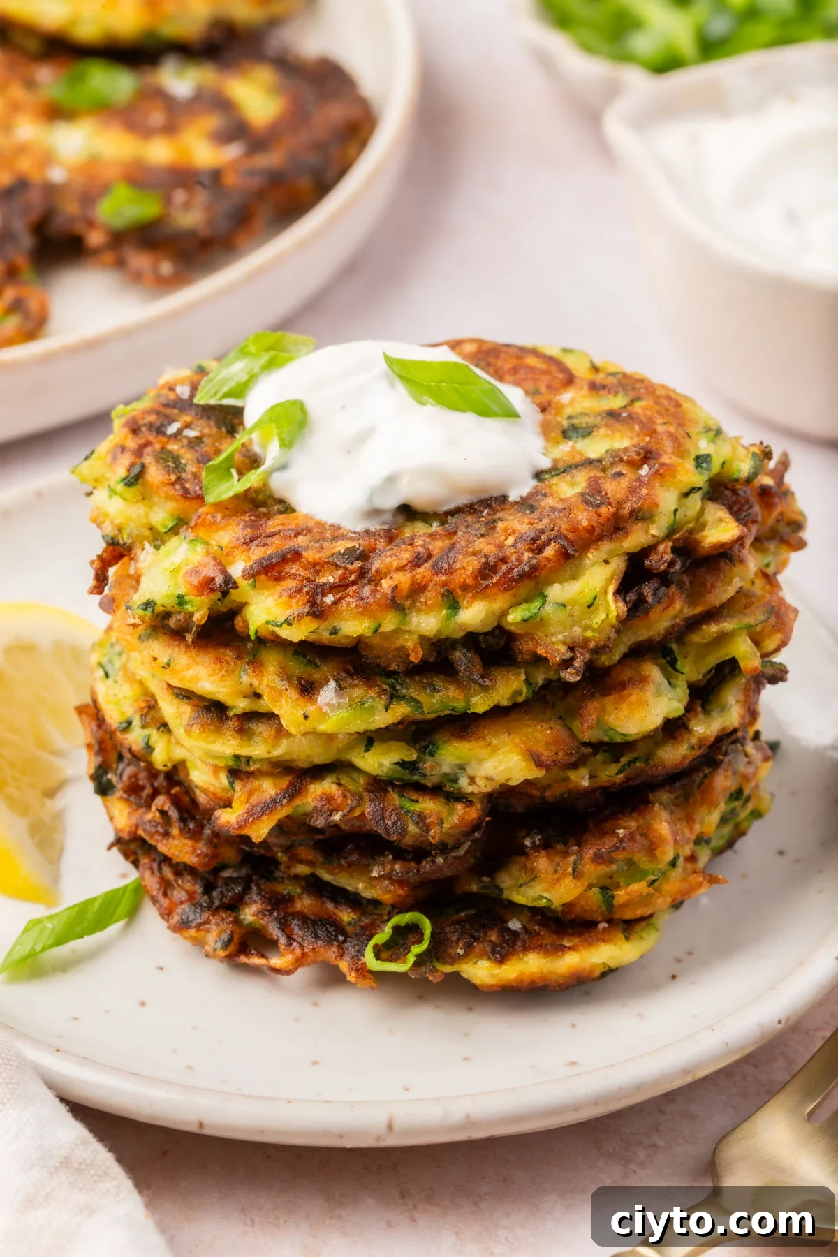Stack of crispy gluten-free zucchini fritters served on a white plate, topped with sour cream and sliced green onions.