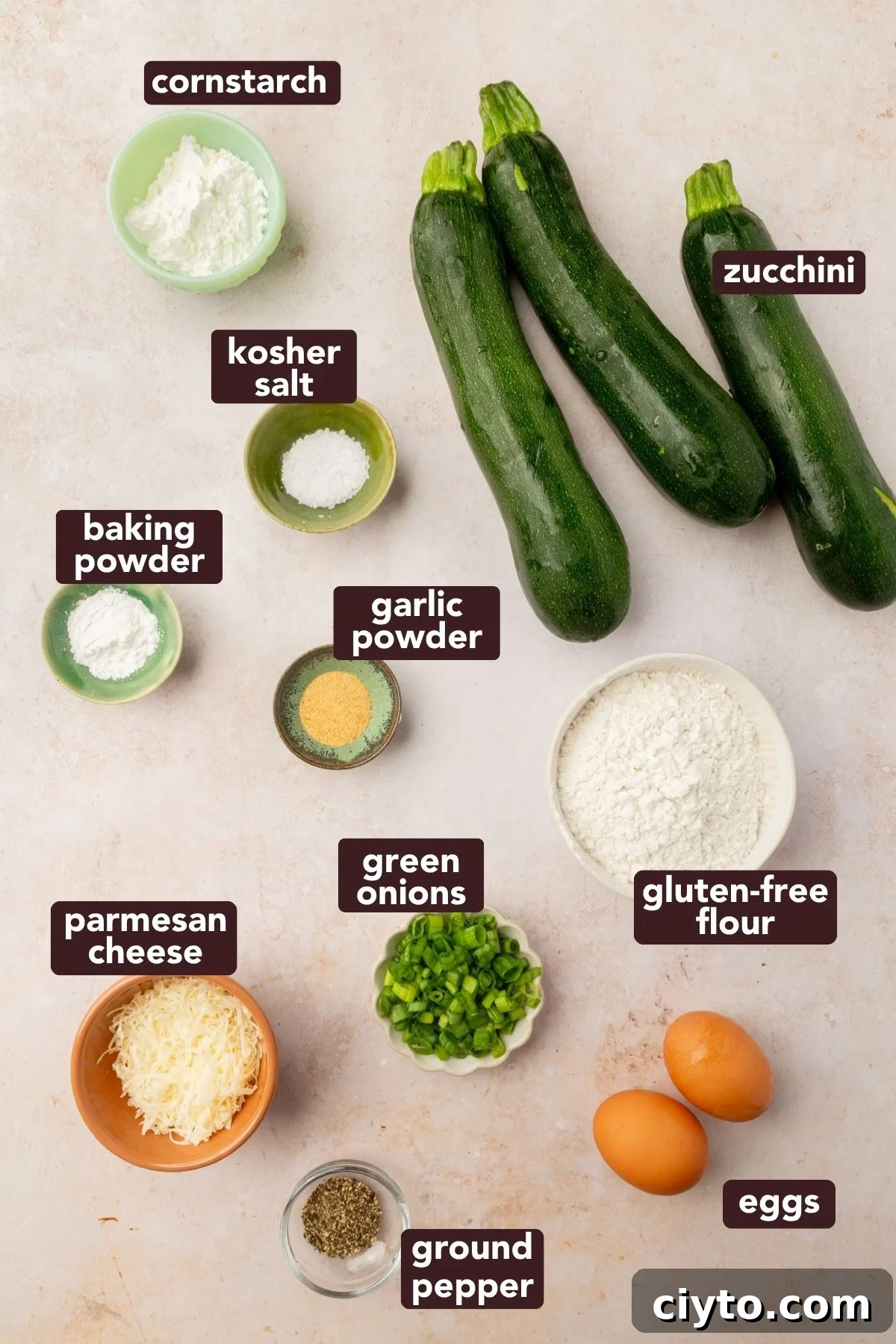 Overhead view of ingredients for gluten-free zucchini fritters, including fresh zucchini, gluten-free flour, green onions, eggs, parmesan, and spices.