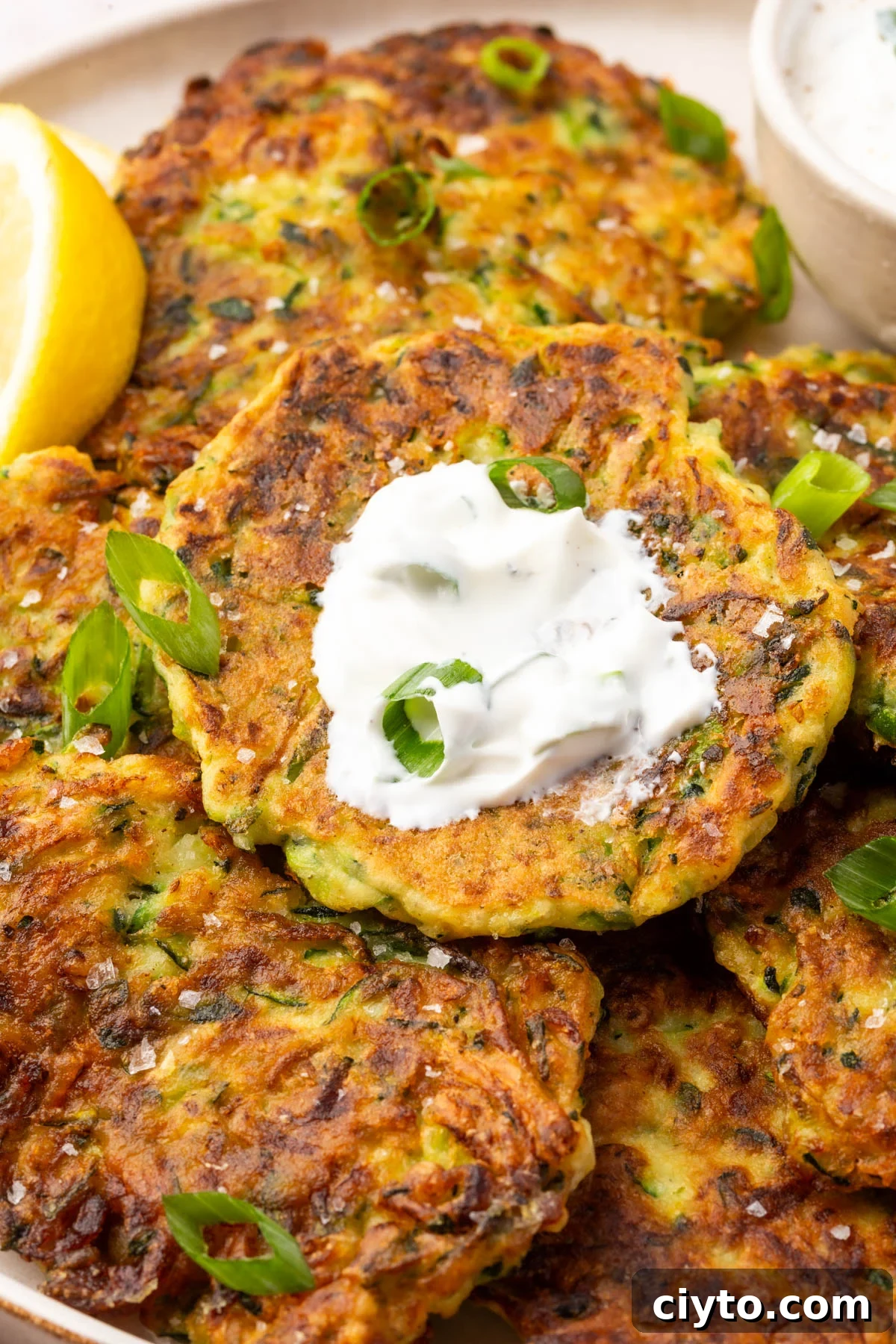 Close-up of a golden-brown zucchini fritter with crispy edges, topped with sour cream and fresh green onions.