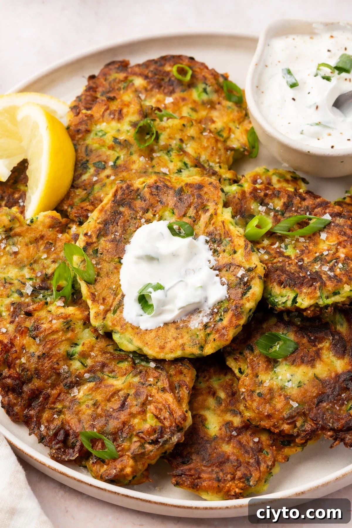 Gluten-free zucchini fritters topped with a dollop of sour cream and garnished with green onion, served with lemon slices and creamy dip on the side.