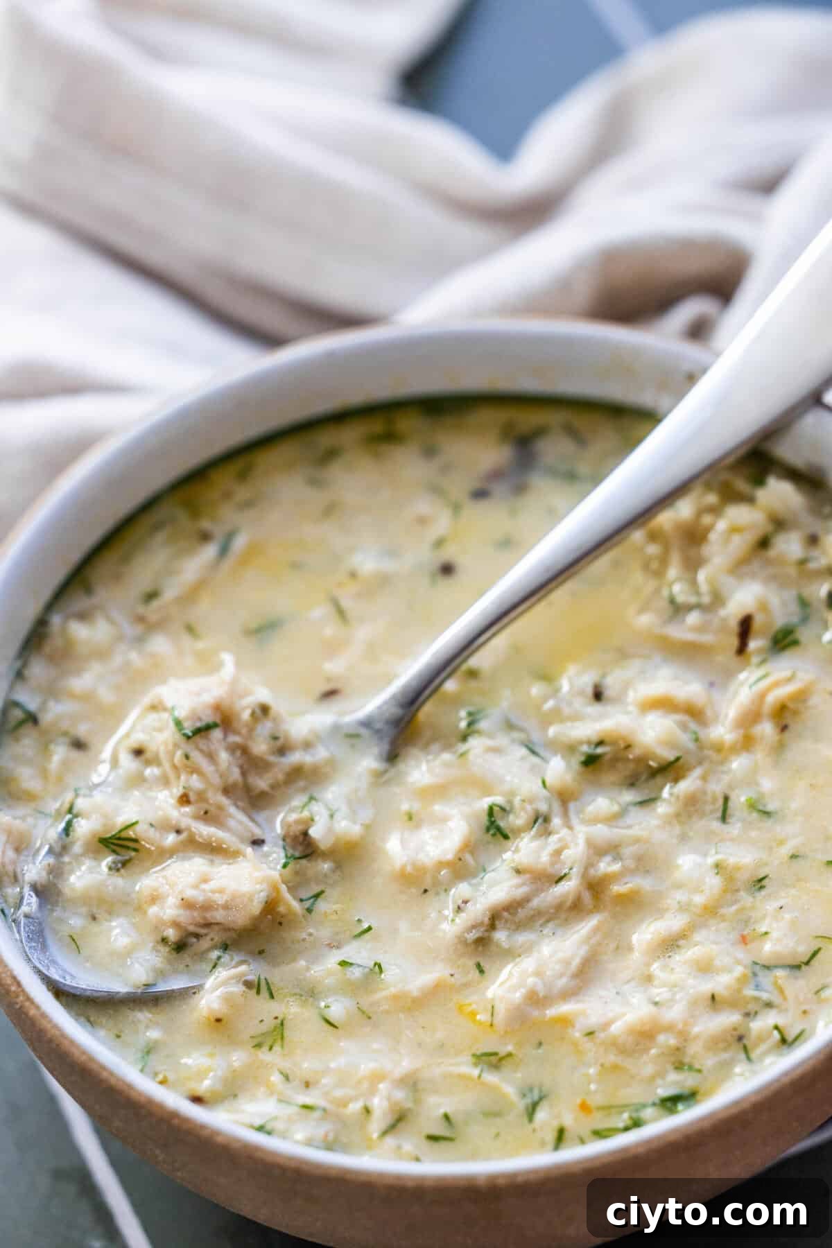 Authentic Greek Avgolemono Soup Recipe 2 A bowl of creamy Greek avgolemono soup with herbs, shredded chicken, and a spoon resting inside. A beige cloth is visible in the background.