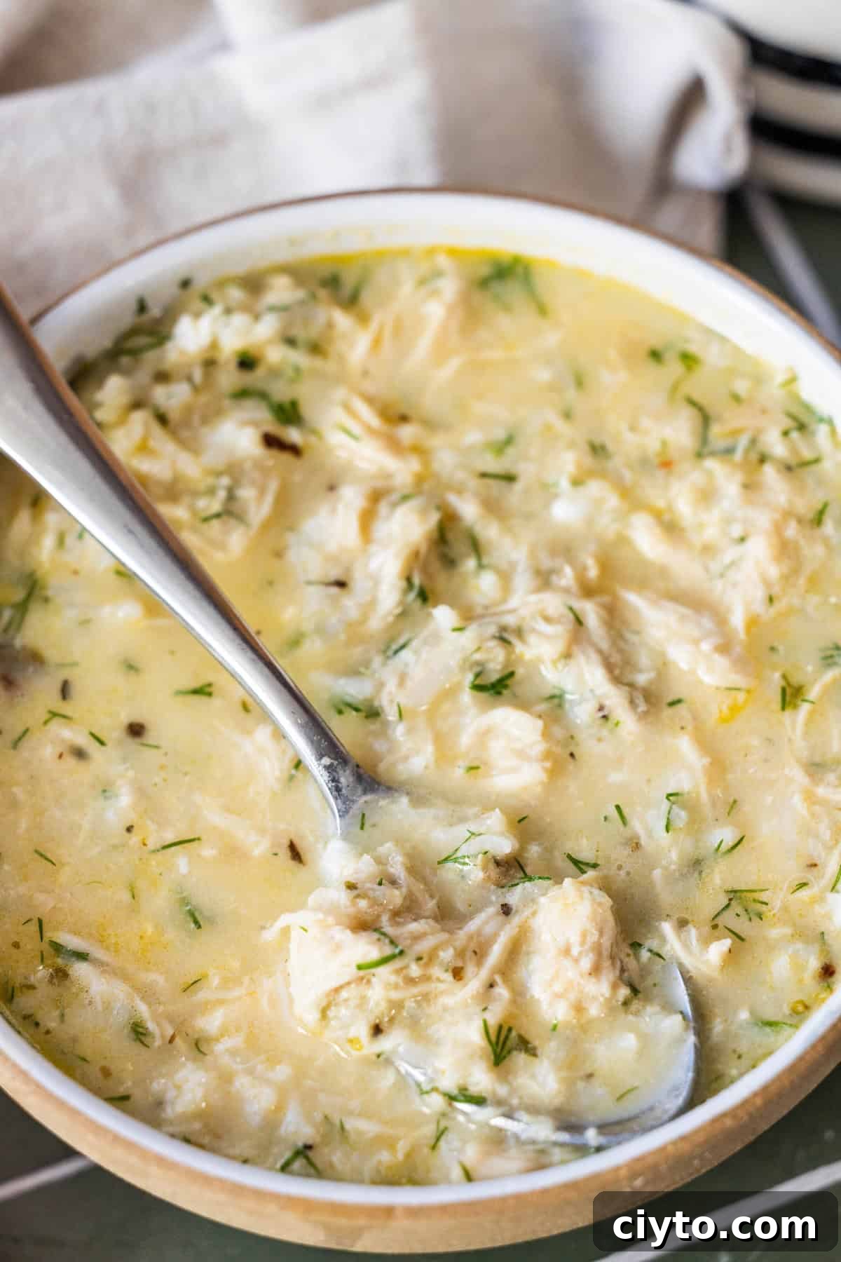 Authentic Greek Avgolemono Soup Recipe 4 A bowl of creamy Greek avgolemono soup with herbs, featuring shredded chicken and a spoon resting inside the bowl.